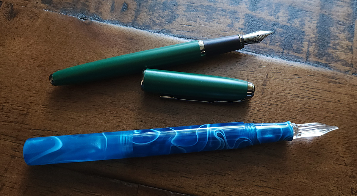 An uncapped dark green fountain pen with a black grip and dark metal clip and a swirly blue resin pen with a clear glass dip nib.