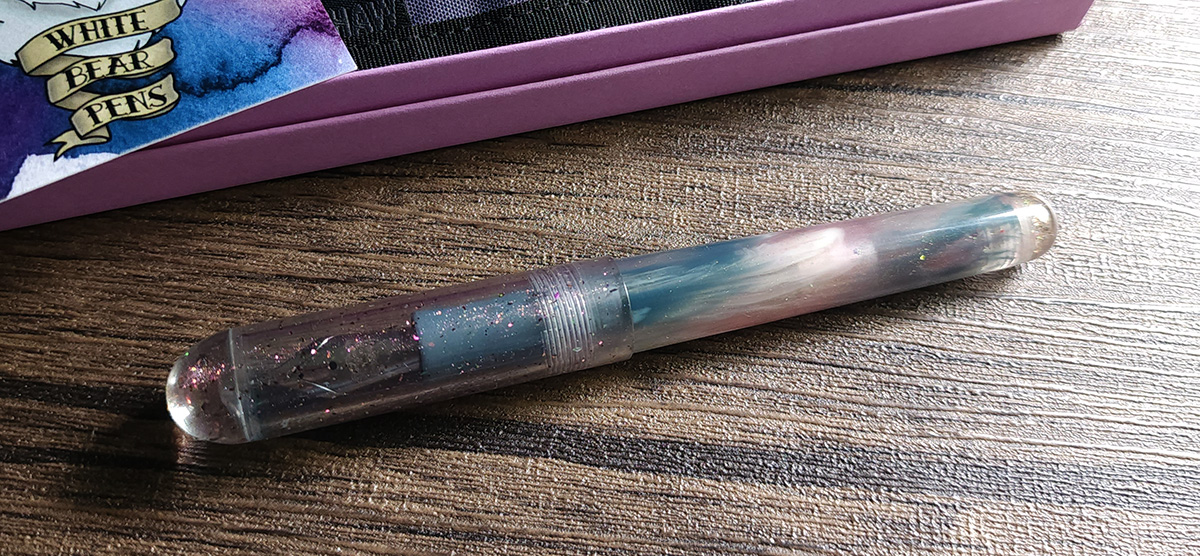 A resin fountain pen with clear dome cap filled with orange/pink glitter and soft blue gray, ivory, and dusty pink body.