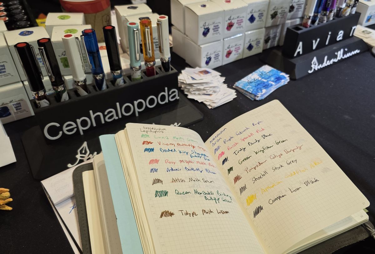 An open A6 notebook with writing tests of the Lepidoptera and Avian series of eight inks each at the Anderillium table with TWSBI Eco tester pens inked up with each ink and ink bottles available for sale.