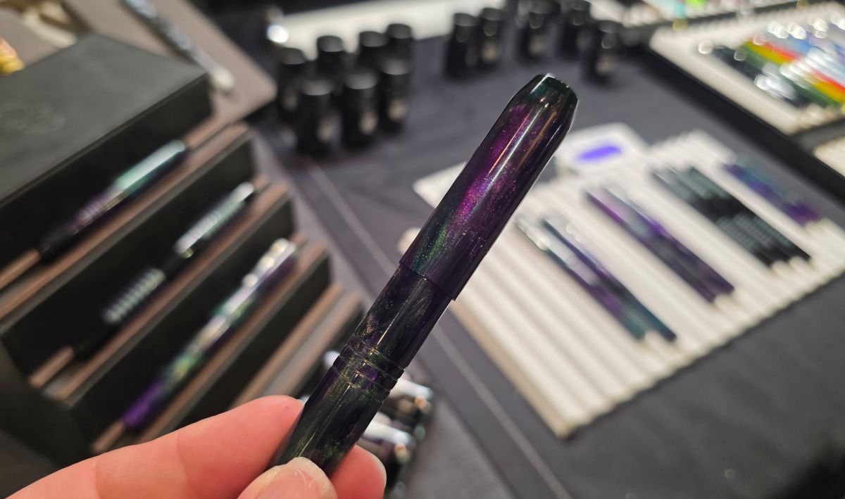 Another small Franklin-Christoph fountain pen in purple and green held up in front of the Franklin-Christoph table at the pen show with more matching pens and ink out of focus in the background.