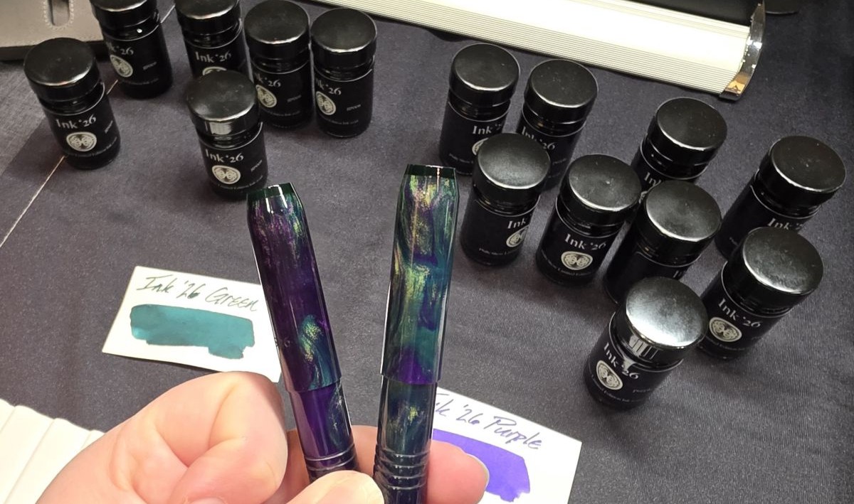 Two small Franklin-Christoph fountain pens in swirled purple and green material held up in front of matching green and purple fountain pen inks at the Franklin-Christoph table.