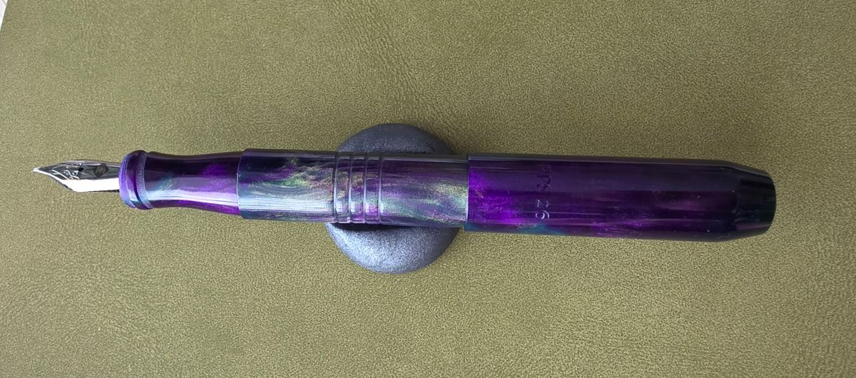 Vibrant purple and swirled green pocket size Franklin-Christoph fountain pen, uncapped with the cap posted on the back, in a clay pen rest on an olive green notebook cover.