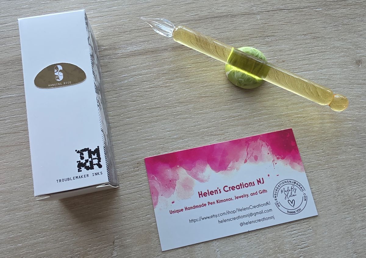 A box of Troublemaker Hanging Rice ink and a short, yellow glass dip pen with a straight handle and ball at the end next to a business card for Helen's Creations NJ.