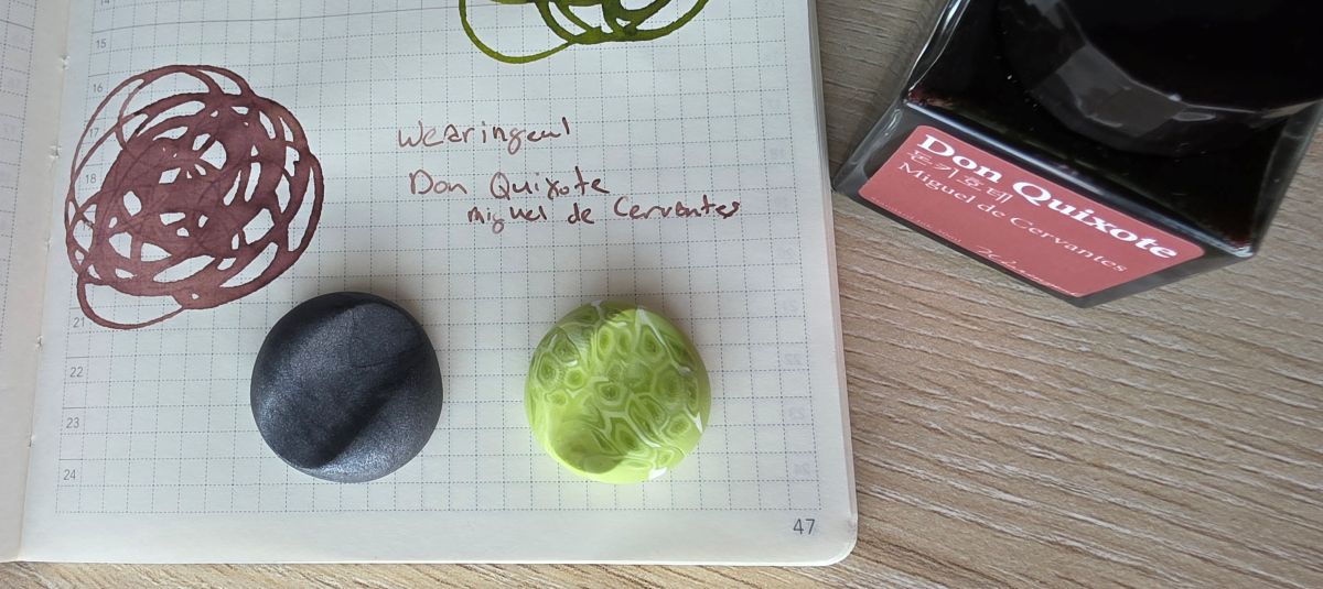 Swatch and ink bottle of Wearingeul Don Quixote, a raisin color ink, and two pen holders made of clay in black and light green with white.