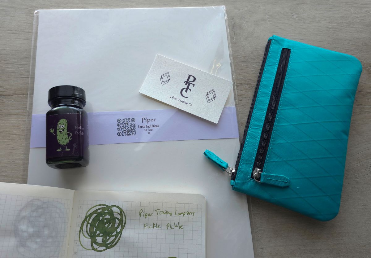 A4 plain paper from Piper, swatch and bottle for the green Fickle Pickle fountain pen ink, a bright teal X-Pac zipper pouch, and business card for Piper Trading Company.
