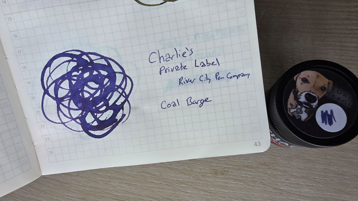 Swatch of Charlie's Private Label Coal Barge ink from River City Pen Company, a dark purple blue shade, next to the bottle of the ink.