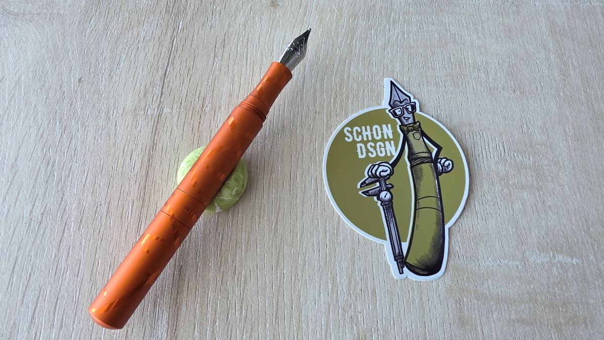 A dark orange matte with shiny lines Schon Dsgn Pocket Six fountain pen, uncapped with a broad nib, next to an olive green Schon Dsgn sticker.