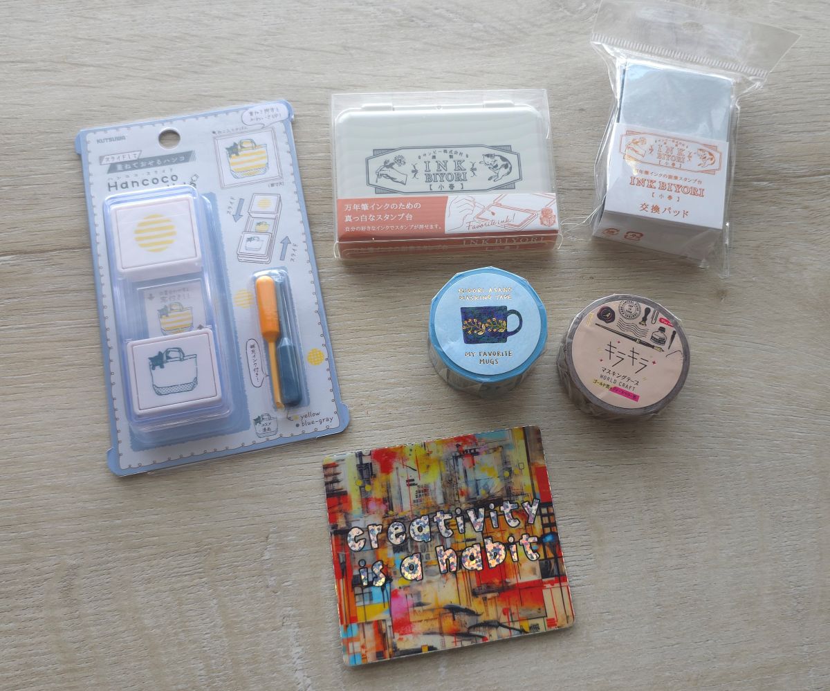 A double-layer stamp set with yellow and blue gray ink and black cat in a tote bag design, an empty ink pad and refills that can be inked with fountain pen ink, two rolls of washing including a mugs design and a red, black, and brown stationery items design, and vinyl sticker with the words 'Creativity is a habit' over a painted background.