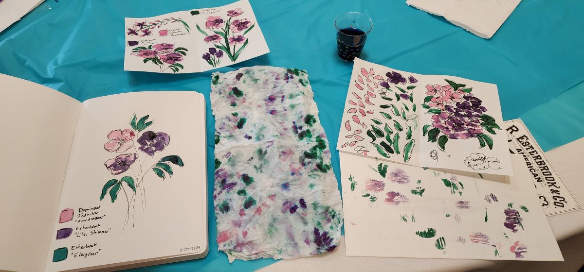 Table during abstract flower painting class showing practice sheets with shapes and floral bouquets in green, pink, and purple ink, 