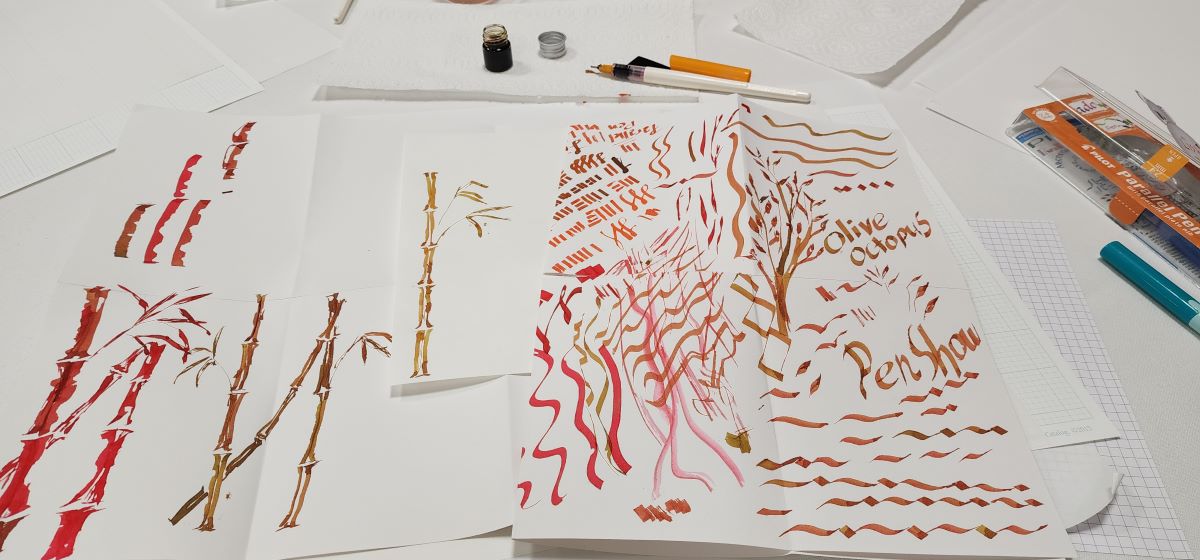 Table during parallel pen class with a Pilot parallel pen, small open bottle of ink, and test sheets with lots of lines, shapes, words, and bamboo.