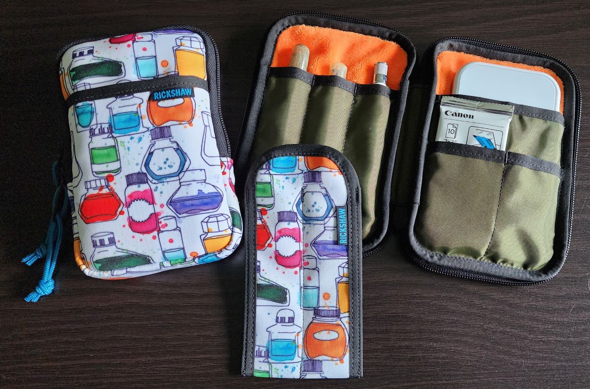 A pen pouch and two pen sleeve in a printed fabric with multi-colored ink bottles, and an open pen case in olive and orange with pen slots on one side and slip pockets on the other.