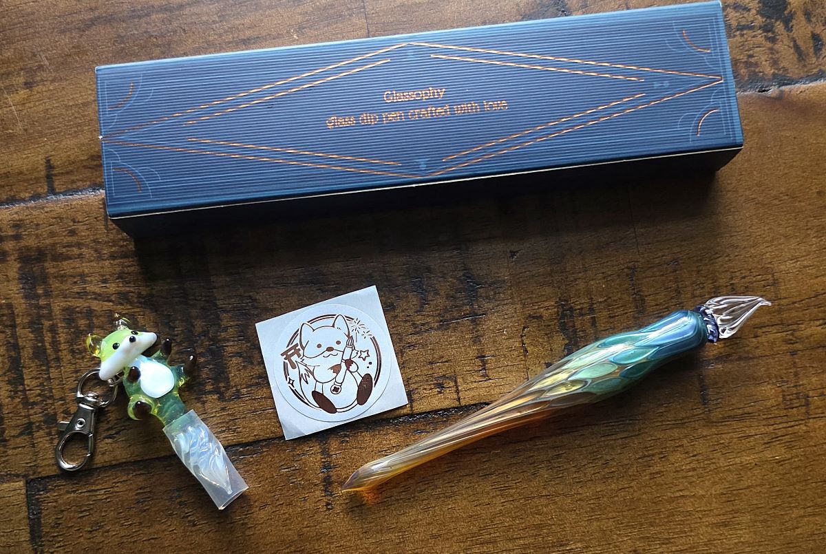 A small green fox made out of glass with a pen nib tail, multicolor glass pen with fude-style tip, sticker of fox holding a pen, and pen box that says Glassophy, Glass dip pen created with love.