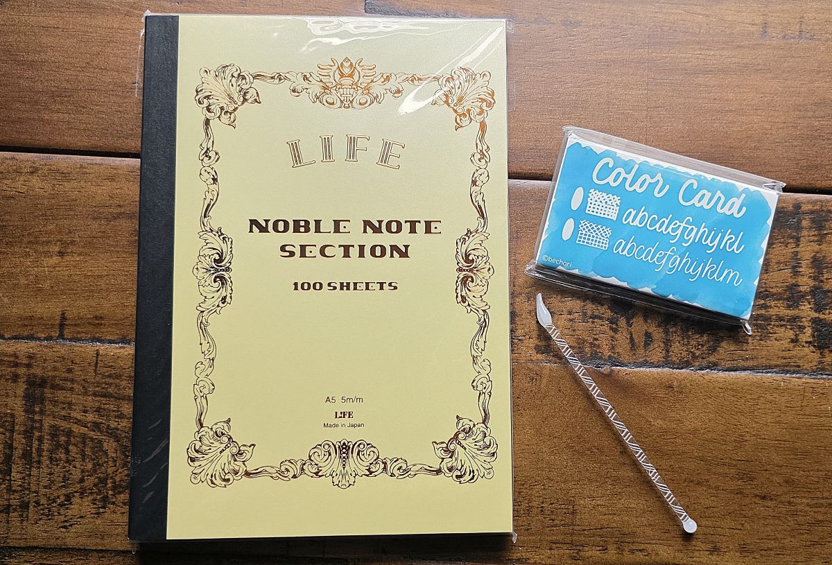A5 Life Noble Note Section notebook with yellow cover and metallic foil design, pack of ink resist swatching cards, and small glass swatching tool with one curved end and a ball on the other end.