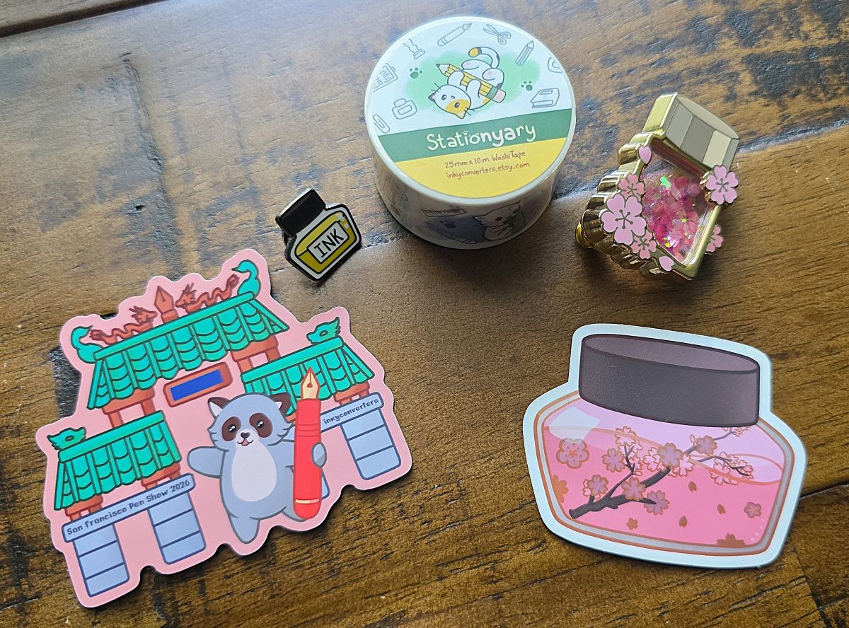 A roll of Stationyary washi tape with cartoon cats, 3D enamel sakura pin with floating blossoms inside, small yellow ink bottle pin, stickers with sakura ink bottle and SF pen show sticker.