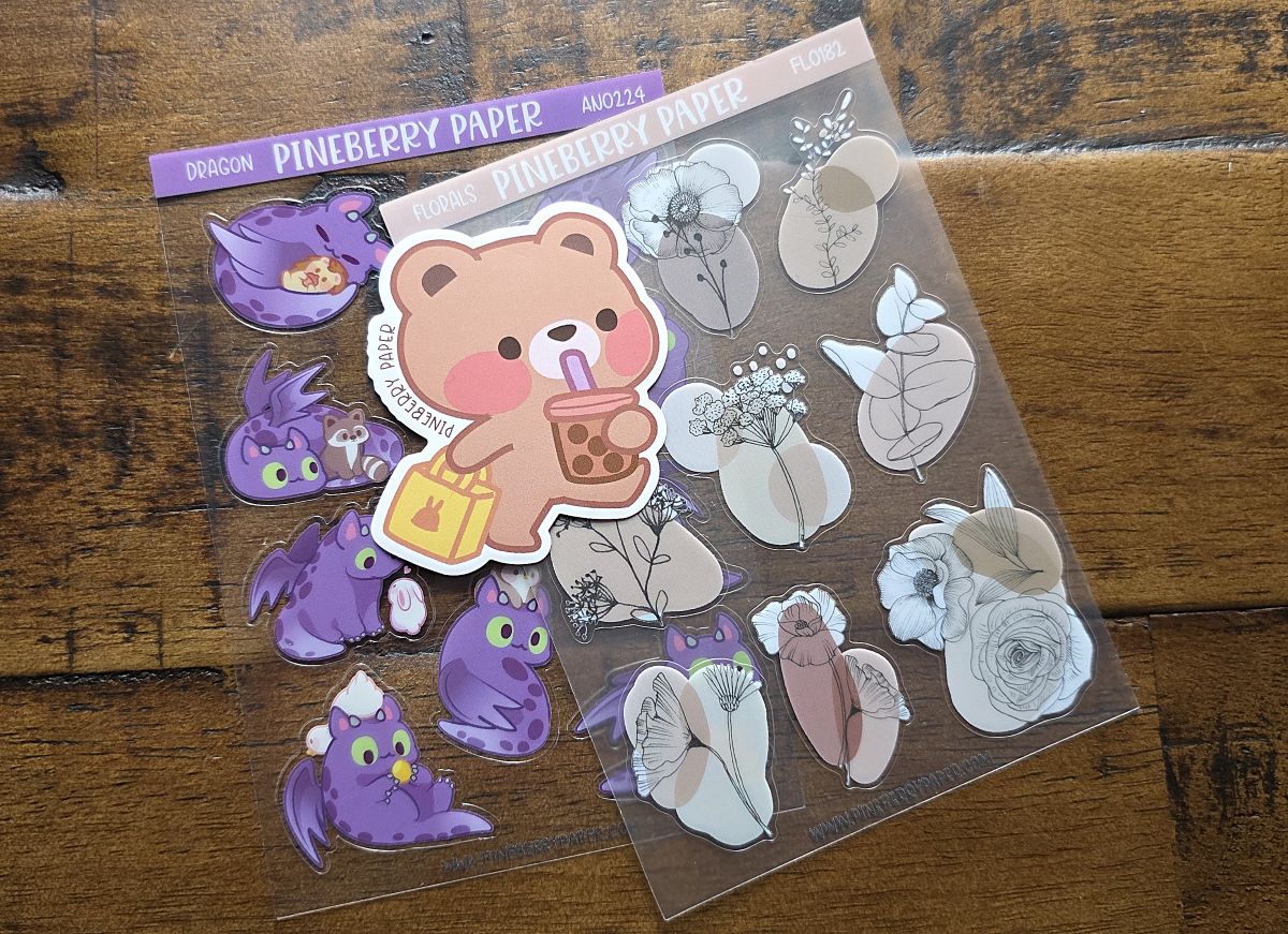 Two sheets of stickers of cute purple dragons and neutral florals plus a vinyl sticker of a teddy bear drinking boba tea and carrying a shopping bag, all from Pineberry Paper.