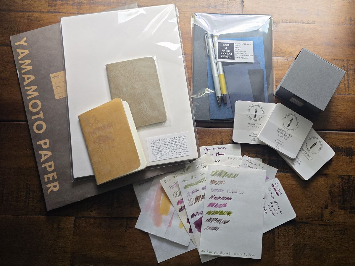 Looseleaf paper, black correspondence paper with 2 metallic Pilot Juice pens, a small box with square sample paper packs, two Ro-biki pocket notebooks with translucent covers, stack of papers with writing and ink tests.