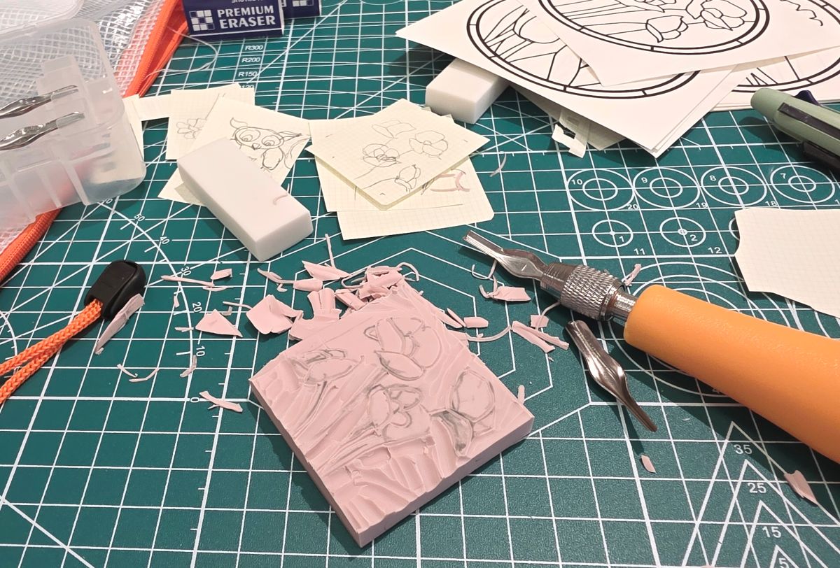 Initial stages of carving a rubber stamp with a carving tool on a cutting mat covered in sketches and shavings.