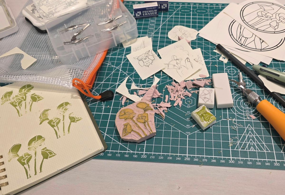 Two completed carved stamps of flowers and a tiny owl in the midst of sketches, shavings, carving tool and pencils, additional erasers to carve, and notebook with test stamp impressions.