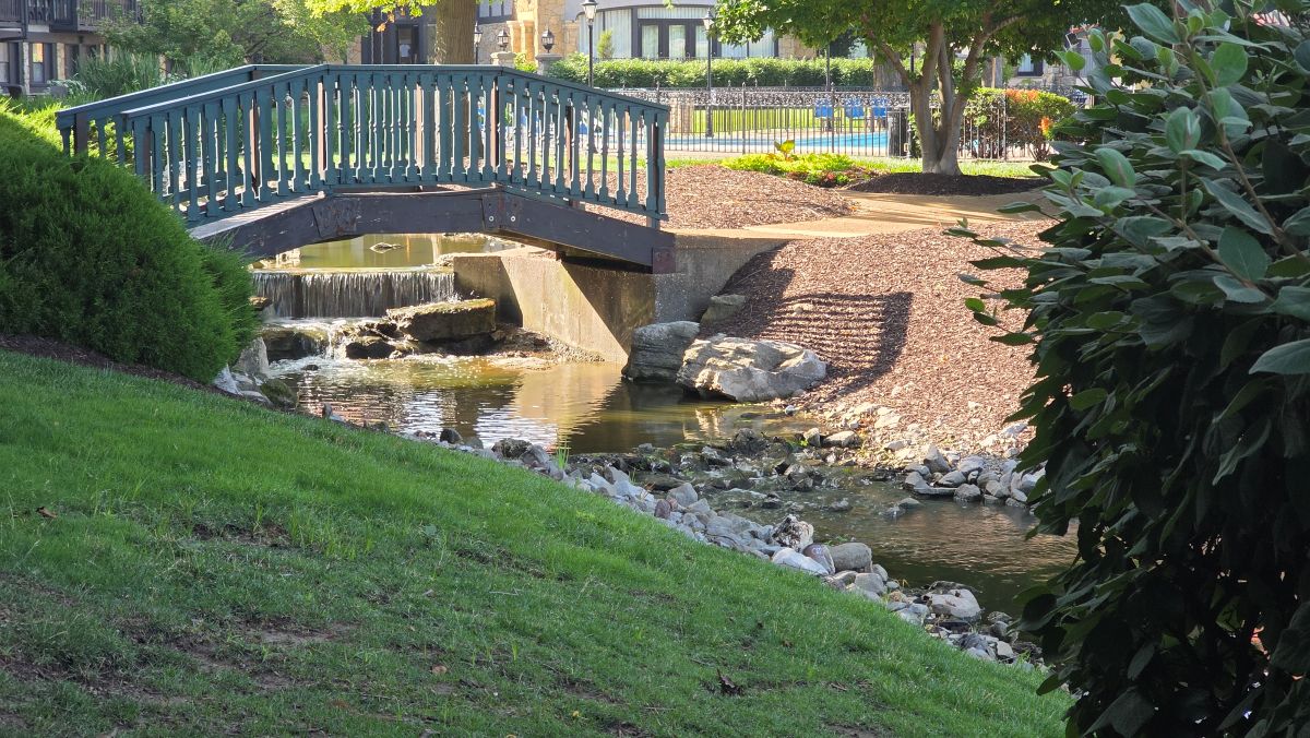 A small bridge for the walking path over a narrow passage of water with a small waterfall surrounded by nice landscaping.