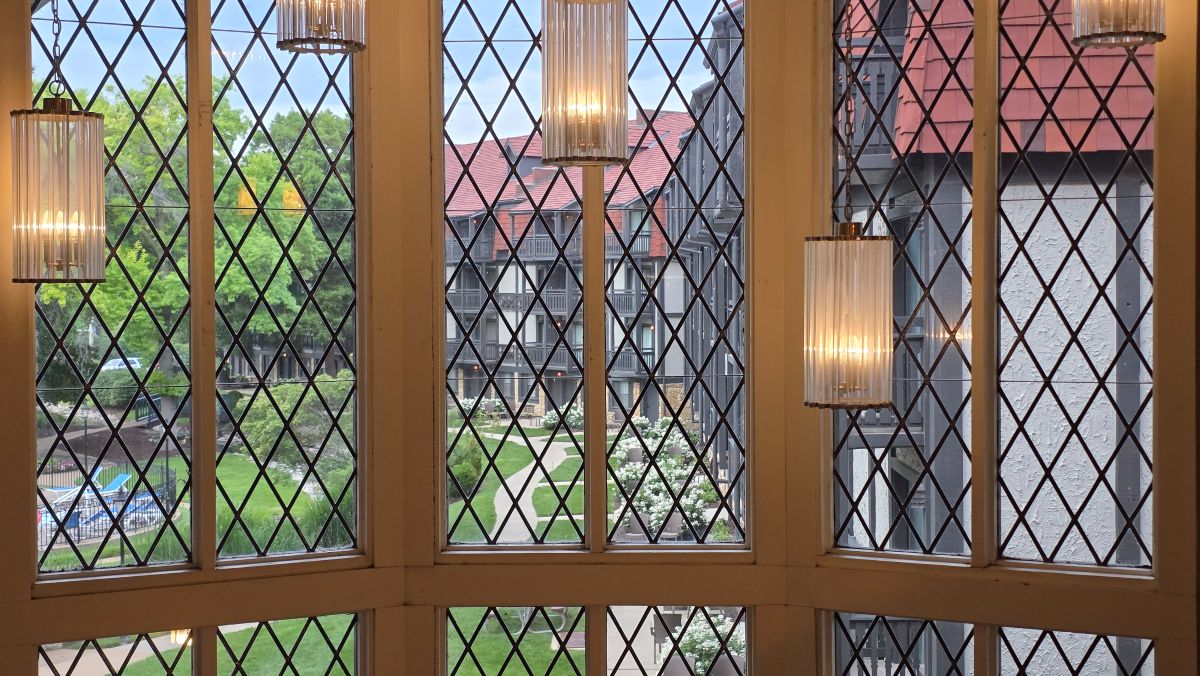 A large set of windows with diamond pattern leading and the back side of the hotel visible outside with a walking path through green grass and trees.