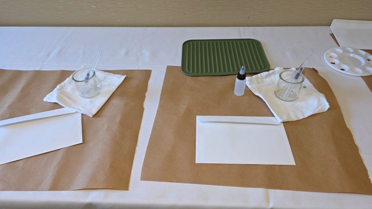 Table setup at the ink painting seminar with an envelope of practice sheets and painting tools including a water brush, glass stir rod, small glass rinse jar, white flour sack towel, and small squeeze bottle of water.
