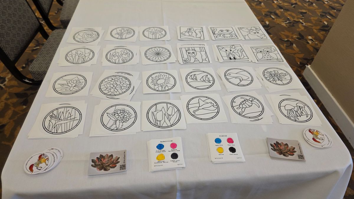 Table with multiple stained glass designs printed out for painting, stickers, and CMYK ink palette swatch sheets for Diamine and Anderillium inks.