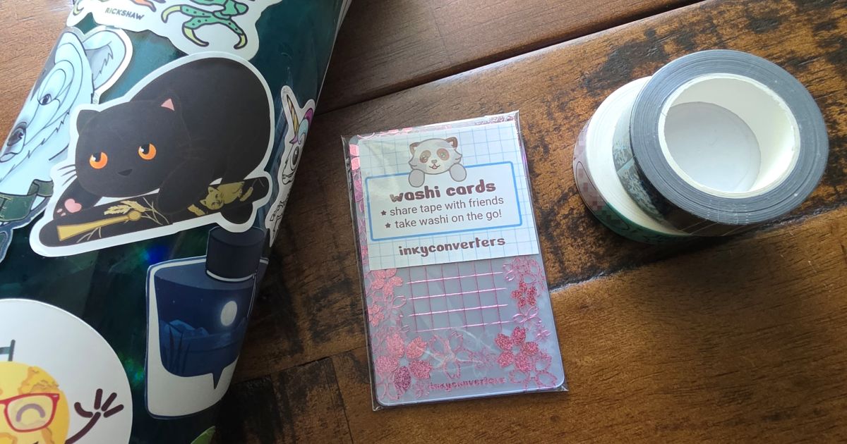 A black cat holding a Maki-e pen sticker on a water bottle, pack of washi cards with pink Sakura design, and two rolls of washi tape.