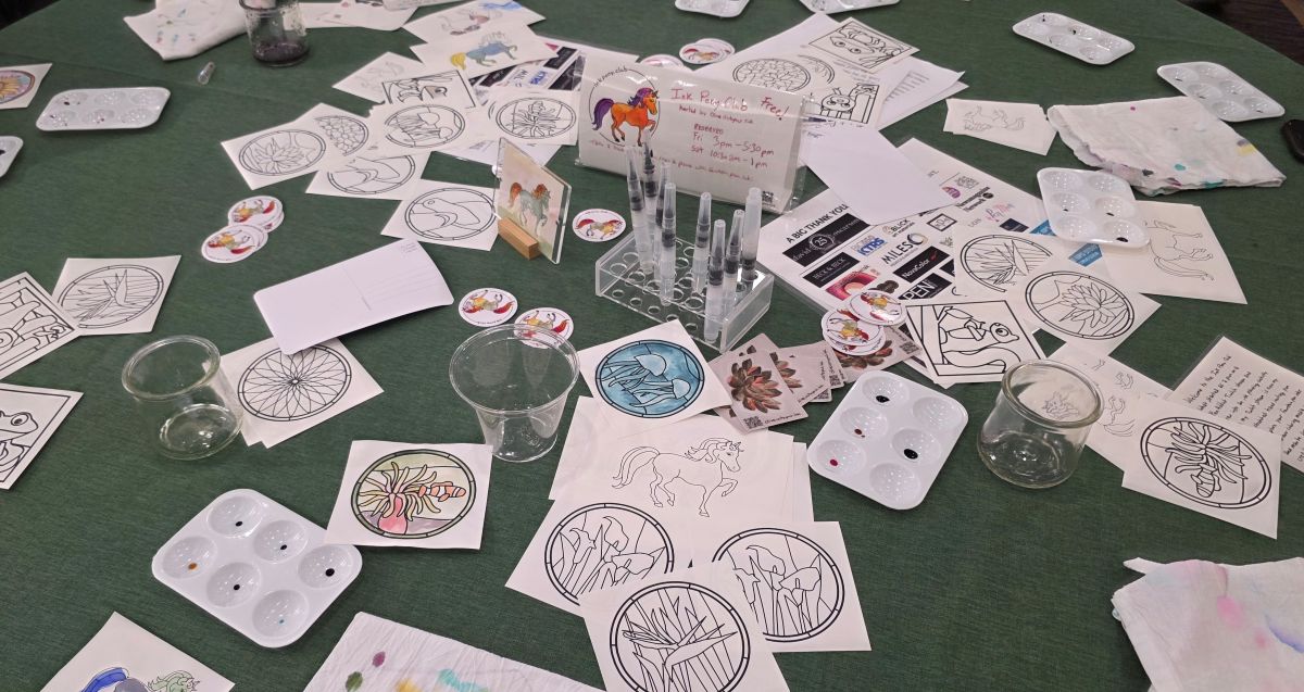 A large round table covered in a dark green table cloth and lots of designs printed on pieces of paper to paint, waterbrushes, and paint palettes with drops of fountain pen ink.