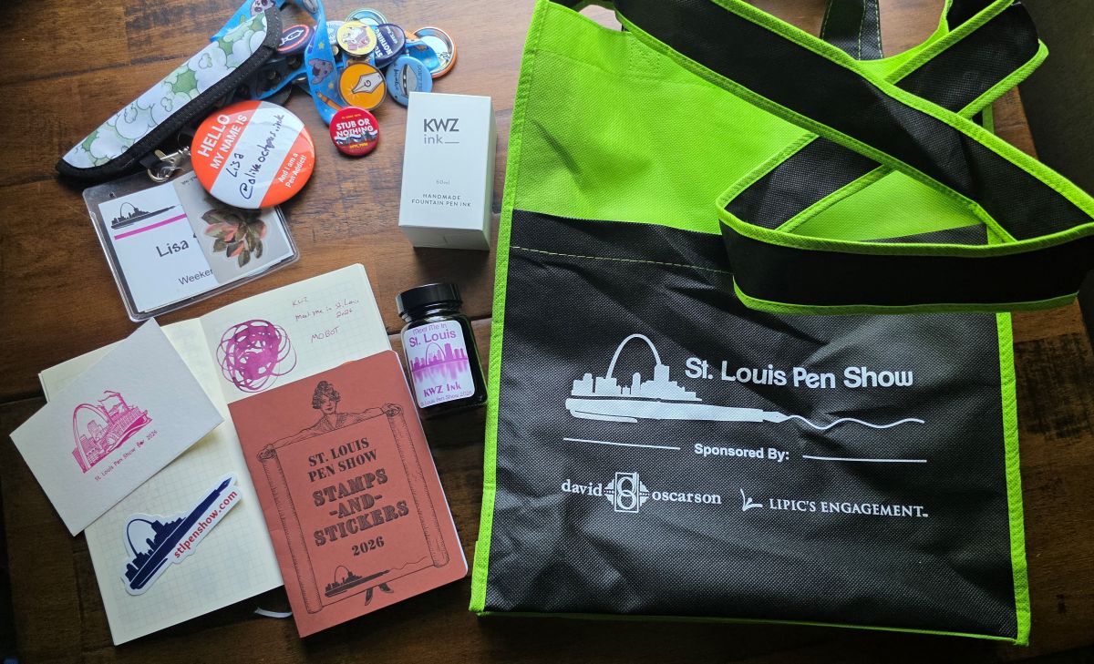 A large green and black sling bag with pen show and sponsor logos, a large bottle of KWZ St. Louis show ink and ink swatch, an A6 size notebook for stamps and stickers, a small sheet of blotter paper with a letter press imprint of the St. Louis Skyline in pink, a pen show logo sticker, and a pen show lanyard covered in pins, with nametag and pen sleeve.