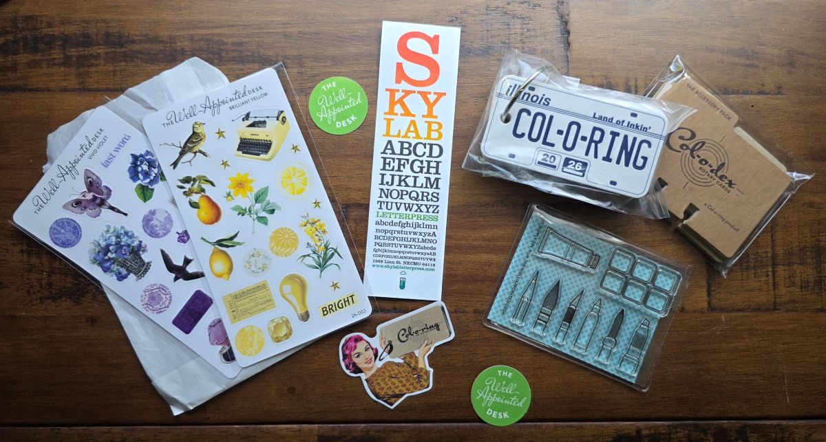 Two sticker sheets with color themes in purple and yellow, a Chicago licence plate themed Col-o-ring, Col-o-dex index tabs, stamps with a paint palette and various art supplies, Skylab Letterpress bookmark and Well-Appointed Desk stickers.