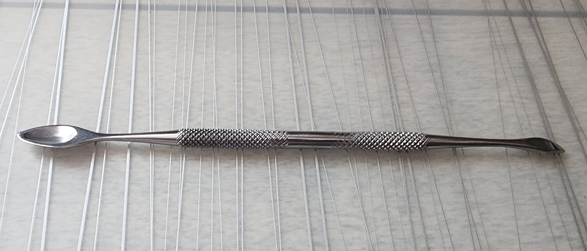 A thin metal hand tool with knurled grips at each end, one end with a small teardrop-shaped spoon and the other with a narrow curve angling down to a point.
