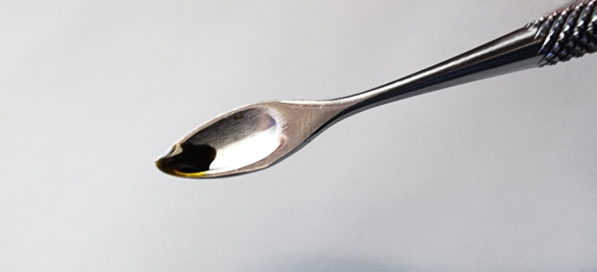 A metal tool with a knurled grip and a small teardrop-shaped spoon at one end holding a drop of ink.