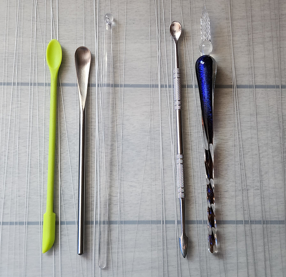 Five other tools placed on a desk, a bright green silicone tool with a narrow spoon on one end and a small scraper tip on other, a metal drink stirrer with an elongated narrow spoon end and and straight circular handle ending in a shallow curve, a clear plastic drink stirrer with a flat stick and small round ball at the end, a metal hand tool with a round spoon at one end and an arrow-shaped point at the other, and a glass dip nib pen with a long twisted glass nib, a bulbous grip, and a tapering twisted handle ending in a narrow rounded tip.