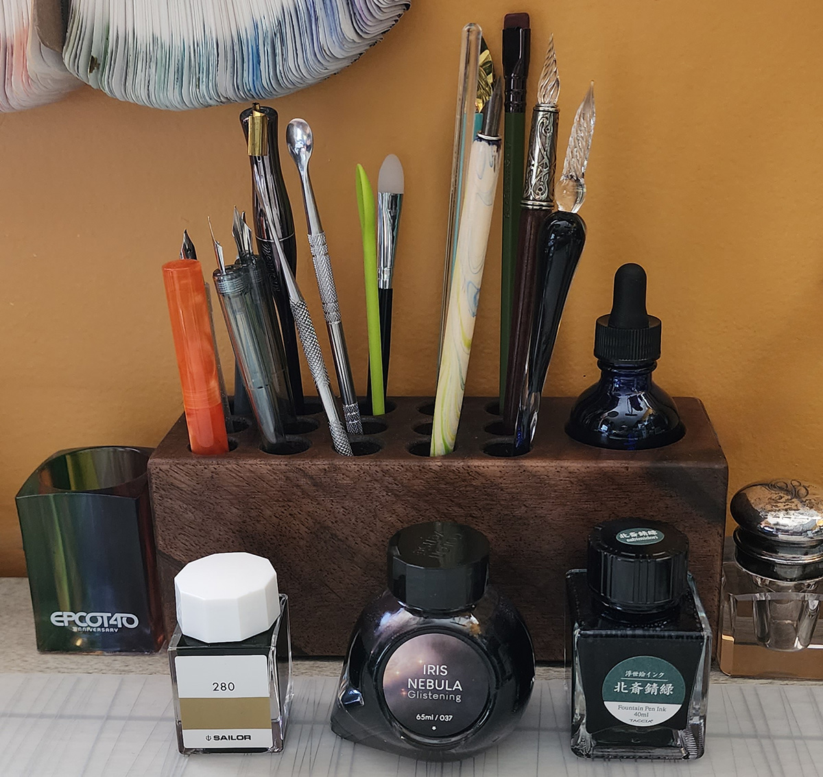 A wooden pen and tool storage block with 15 pen slots filled with a variety of dip pens and swatching tools, and a small bottle of water with a dropper cap in the single large storage slot.