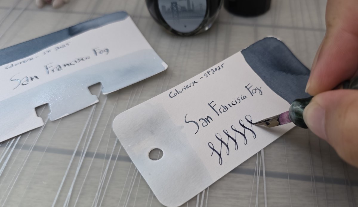 Closeup of a hand writing on a Col-o-ring card with a flexing metal dip nib including the name and maker of the ink, Colorverse San Francisco Fog, and a series of loops with thin upstrokes and wider downstrokes.