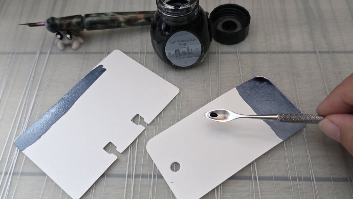 A medium blue gray ink being spread on Col-o-ring and Col-o-dex swatch cards by a tiny metal carving tool shaped like a spoon that has been dipped in the ink, with the open ink bottle and dip pen in the background.