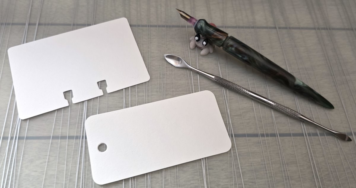Single blank Col-o-ring and Col-o-dex ink swatch cards  laid out for swatching next to a small metal carving tool and a metal calligraphy dip nib in a swirled resin nib holder.