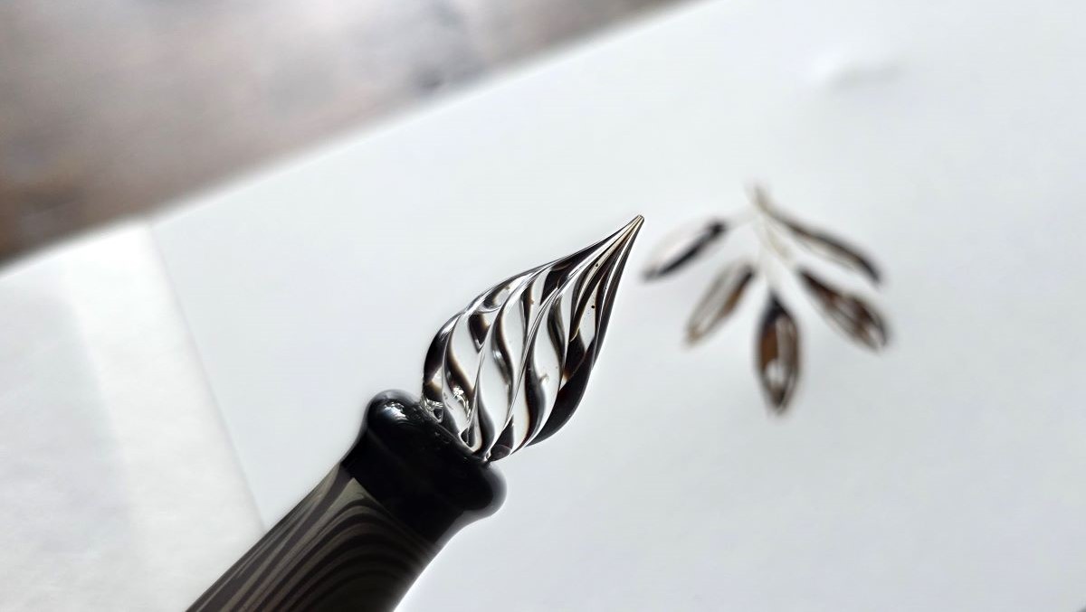 Closeup of the nib of a Hanabi glass pen with dark ink in the channels and a sketch of dark leaves out of focus in the background.
