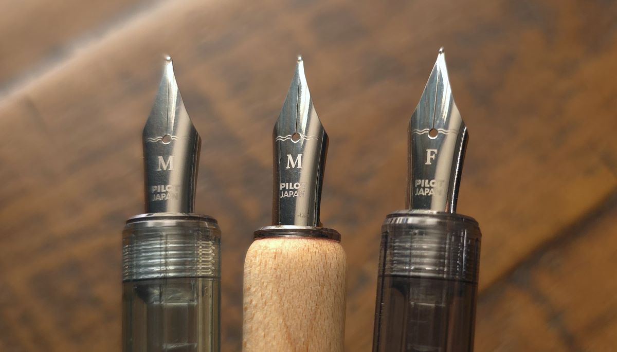 Closeup of the nibs of 3 Pilot Iro-Utsushi dip pens: a medium nib with a translucent pale blue handle, a medium nib with a light wood handle, and a fine nib with a translucent dark gray handle.