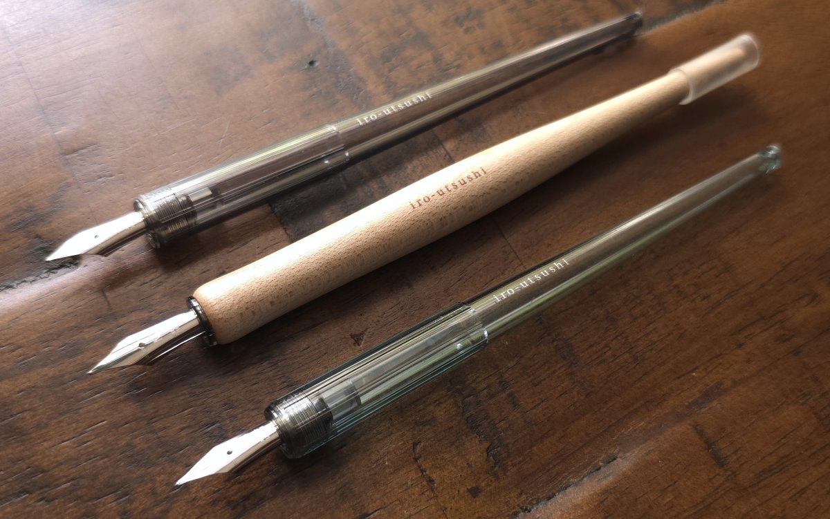 Three Pilot Iro-Utsushi dip pens on a desk: one with a translucent dark gray handle and a fine nib, one with a light wood handle and medium nib, and one with a translucent pale blue handle and medium nib.