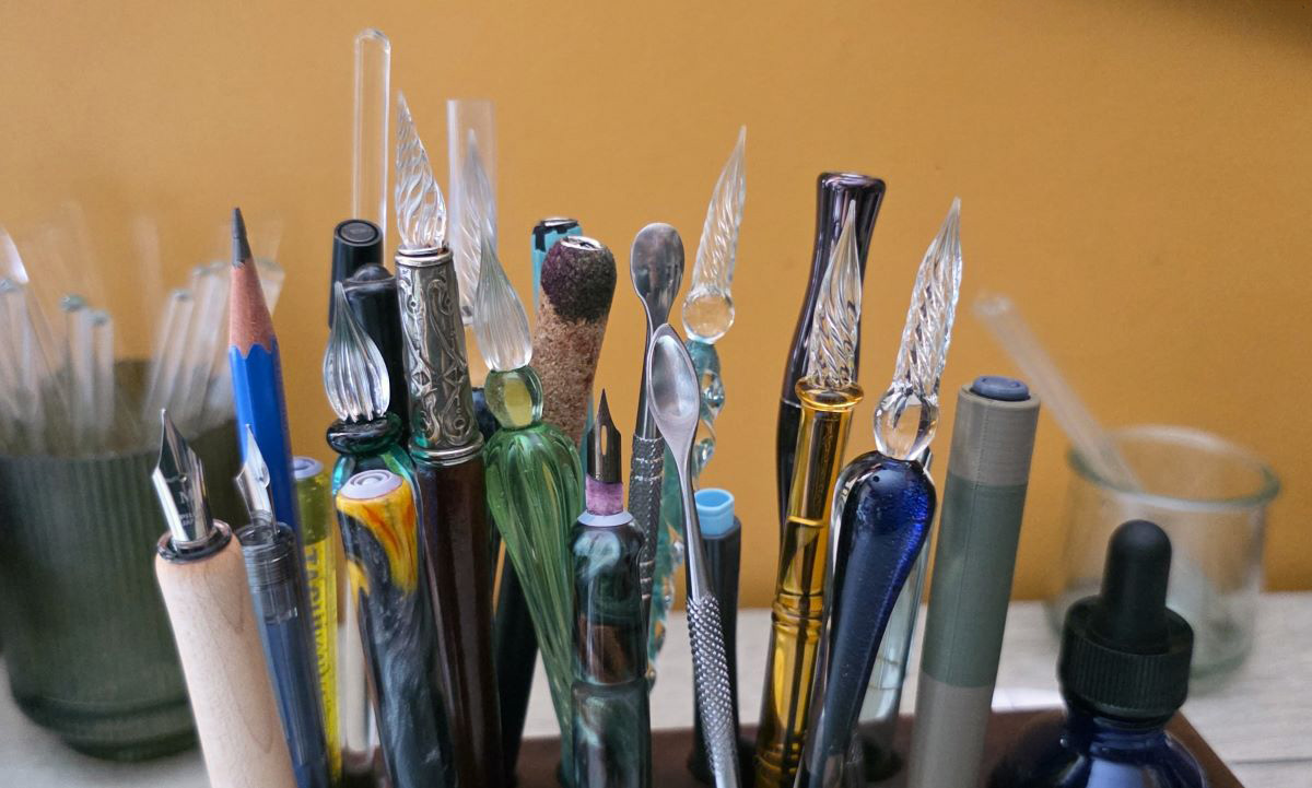A wooden block with 15 slots to store writing tools, filled with glass pens, nib holders, metal dip pens, a long glass stir rod, metal carving spoons, and a pencil.