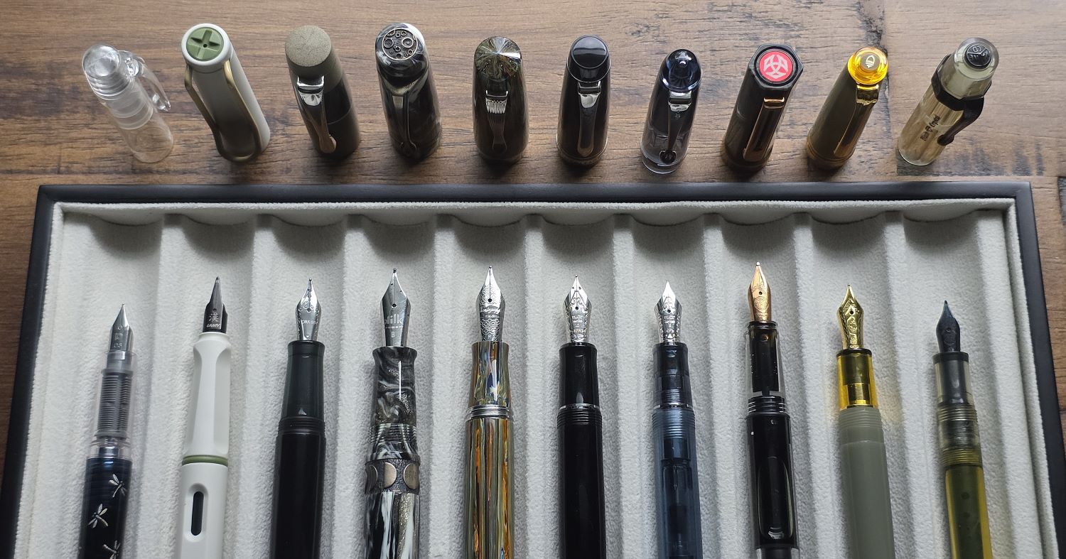 Ten uncapped fountain pens in a lined drawer with the caps sitting nearby.