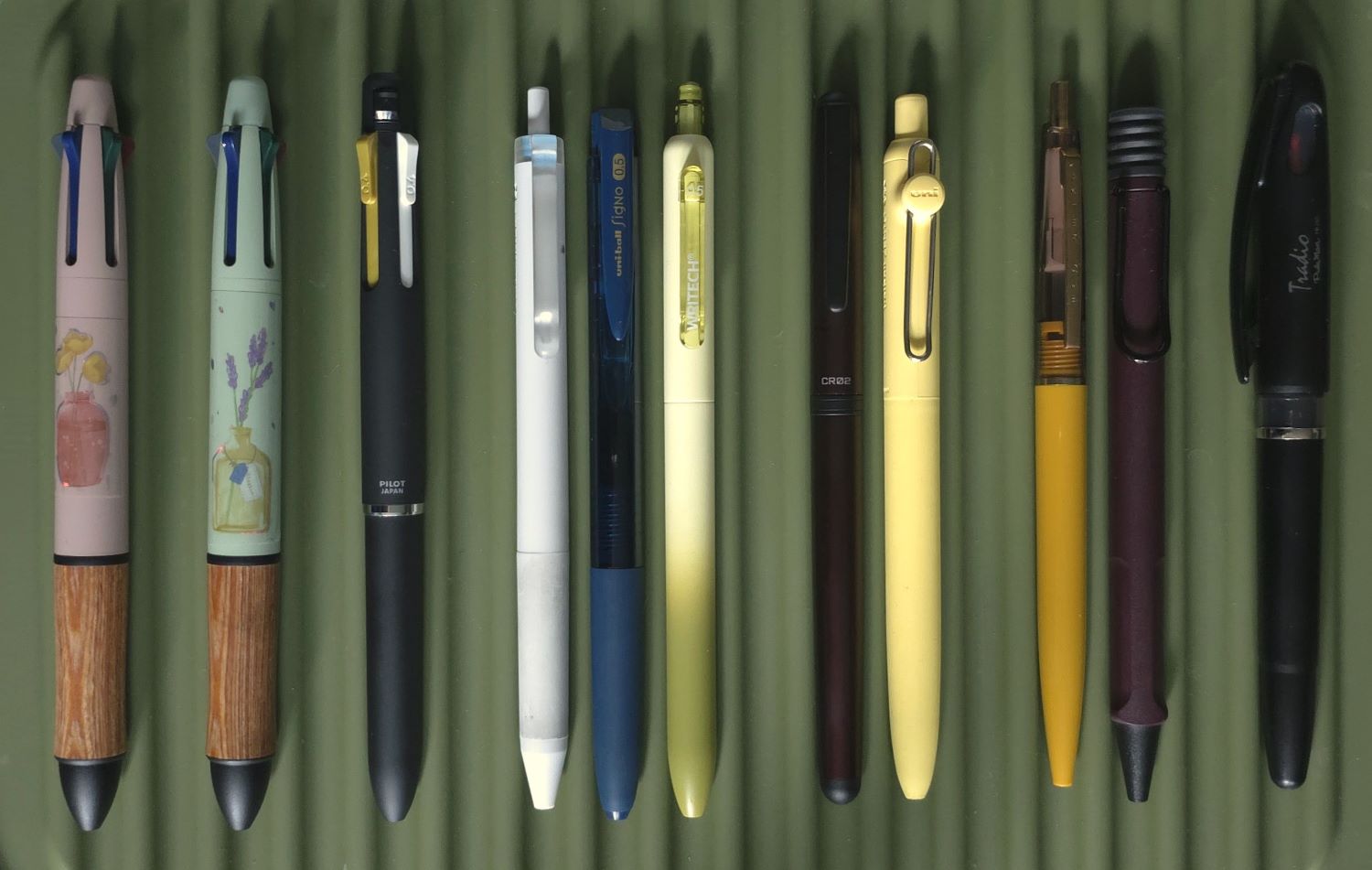 Ten different types of ballpoint, rollerball, gel, fineliner, and multi pens displayed on an olive green mat.