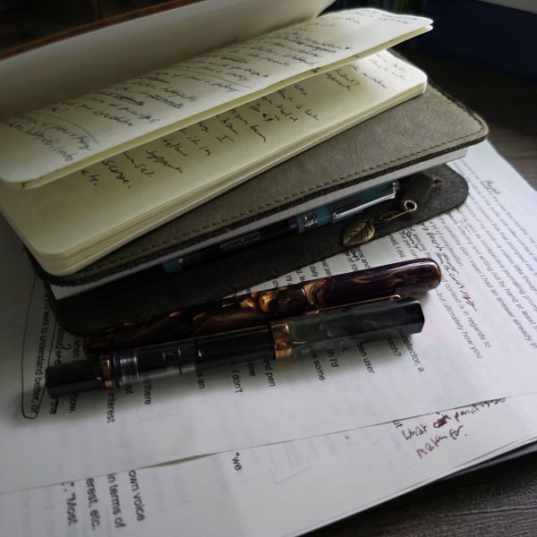 A stack of paper and notebooks including looseleaf pages with printed text and handwritten edits, a green A5 slim Roterfaden Taschenbegleiter filled with notebooks, a Ro-biki notebook, and a couple fountain pens.