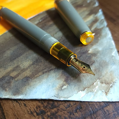 Sailor Pro Gear Slim 'Nuts' pen with a medium gold nib and light gray green translucent body with transparent yellow accents with swatches of the green/brown/blue Sailor Manyo Shirakashi and bright yellow Sailor Manyo Yamabuki inks.