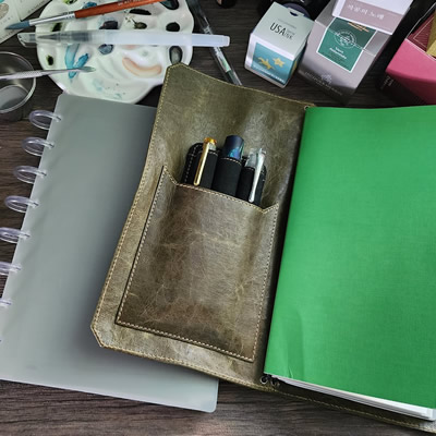 A top-down view of a desk with a Franklin-Christoph TN-size notebook cover with inserts, a 3 pen holder visible in the front cover pocket, on top of a gray TUL discbound Jr notebook with clear disks, surrounded by fountain pen ink boxes and bottles, several dip pens with metal and glass nibs, a paint brush, water brush and ceramic watercolor palette.