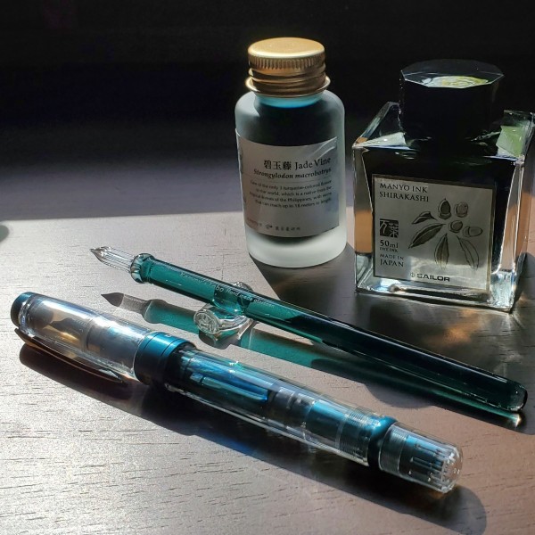 A TWSBI 580ALR Prussian Blue fountain pen, a teal green glass dip pen, and glass bottles of Sailor Manyo Shirakashi and Ink Institute Jade Vine inks on a wood desk.