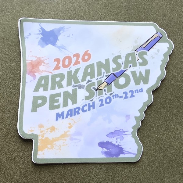 A sticker shaped like the state of Arkansas with ink splotches, an uncapped fountain pen, and the text: 2026 Arkansas Pen Show March 20th - 22nd