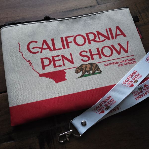 A Rickshaw A5 zipper pouch with the California Pen Show logo design over a cream background with a red block across the bottom, also a wide white lanyard with the California Pen Show logo repeated along the length.