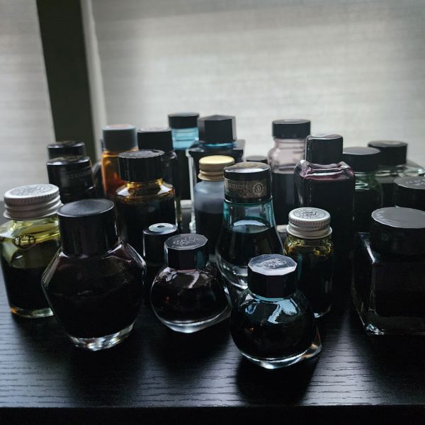 A collection of 24 fountain pen ink bottles on a table, mostly glass but a few plastic in a variety of shapes and sizes, with labels facing away and backlit by light through a white window shade.